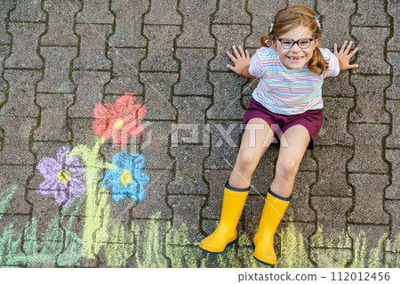 Cute little girl and flowers painted with colorful chalks on asphalt. Happy preschool child having fun with painting chalk picture. Creative leisure for children, drawing and painting. 112012456
