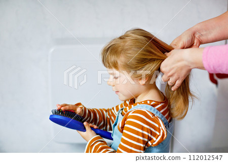 Close up photo of happy smiling toddler girl with blond hair and hands of mum brushing hairs and make plait or pigtail. Mother prepares child for kindergarten or daycare. Family morning routine. Close up photo of happy smiling toddler girl with blond hair and hands of mum brushing hairs and make plait or pigtail. Mother prepares child for kindergarten or daycare. Family morning routine. 112012547