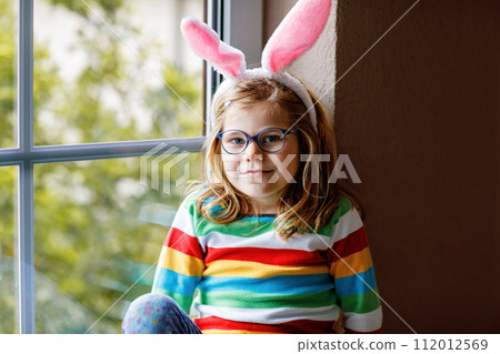 Portrait of a happy little girl with bunny ears looking outside sitting by a window. Easter holiday. Adorable child happy about holiday. 112012569