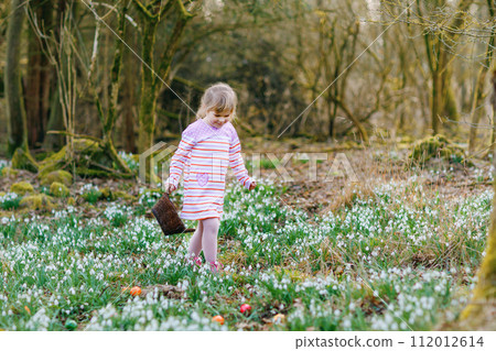 Little girl in pink dress making egg hunt in spring forest on sunny day, outdoors. Cute happy child with lots of snowdrop flowers and colored eggs. Springtime, christian holiday concept. Little girl in pink dress making egg hunt in spring forest on sunny day, outdoors. Cute happy child with lots of snowdrop flowers and colored eggs. Springtime, christian holiday concept. 112012614