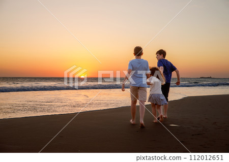 Three kids silhouettes running and jumping on beach at sunset. happy family, two school boys and one little preschool girl. Siblings having fun together. Bonding and family vacation. 112012651
