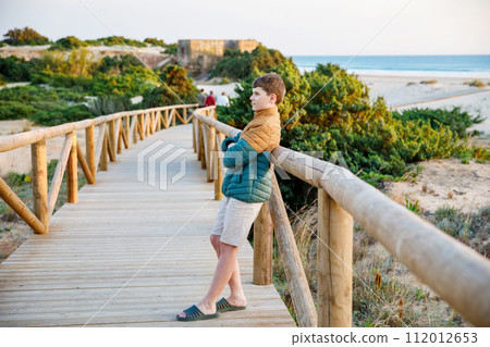 Happy cheerful teenager standing on beach at sunset. happy preteen handsome boy smiling at the camera. Kid on family vacation at the sea. 112012653