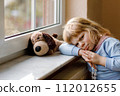 Upset toddler girl sitting by window and looking out. Sad crying child feeling unhappy. Lockdown and isolation concept during corona virus pandemic time. Lonely preschool kid. 112012655
