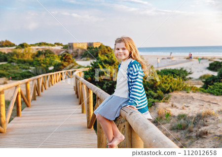 Adorable happy smiling little girl on beach vacation at sunset. Handsome cute preschool child with long blond hairs having fun on family vacations. Adorable happy smiling little girl on beach vacation at sunset. Handsome cute preschool child with long blond hairs having fun on family vacations. 112012658