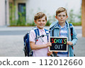 Two little kid boys with backpack or satchel. Schoolkids on the way to school. Healthy children, brothers and best friends outdoors on street leaving home. School's out on chalk desk. Happy siblings. 112012660