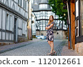 Beautiful young woman with long hairs in summer dress going for a walk in German city. Happy girl enjoying walking in cute small fachwerk town with old houses in Germany. 112012661