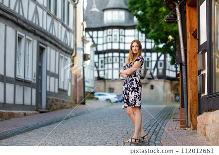Beautiful young woman with long hairs in summer dress going for a walk in German city. Happy girl enjoying walking in cute small fachwerk town with old houses in Germany. 112012661