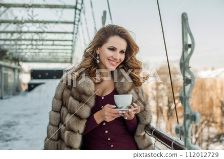 Beautiful brunette woman wearing fur winter coat holding a cup of hot coffee 112012729