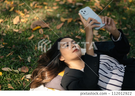 Millennial businesswoman lying on the green grass and typing message on mobile phone Millennial businesswoman lying on the green grass and typing message on mobile phone 112012922