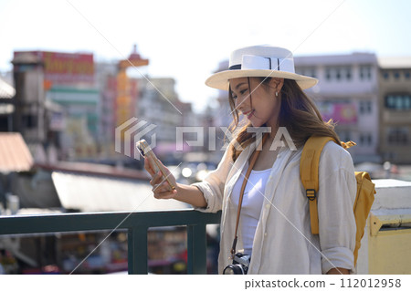 Portrait of smiling female traveller with backpack standing on bridge and using smartphone 112012958