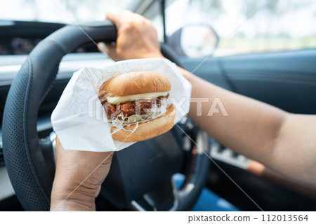 Asian lady holding hamburger to eat in car, dangerous and risk an accident. Asian lady holding hamburger to eat in car, dangerous and risk an accident. 112013564