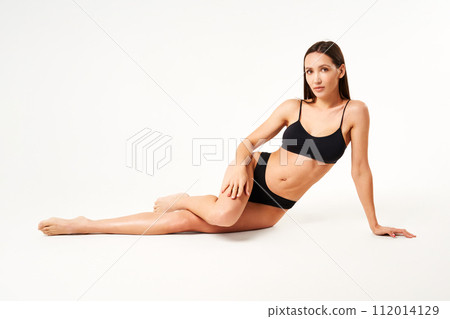 Young beautiful woman with perfect body in black underwear sitting on floor posing on studio 112014129