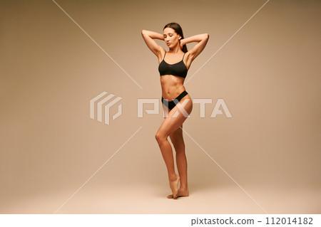 Pretty sporty woman in black underwear with arms up posing on beige background Pretty sporty woman in black underwear with arms up posing on beige background 112014182