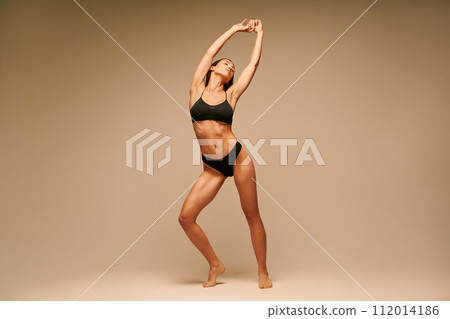 Full length portrait of pretty sporty woman in black underwear with arms up posing on beige background 112014186
