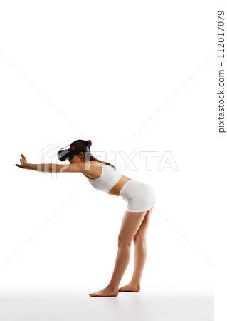 Stretching. Full-length image of young girl with slim body, wearing VR glasses and training against white studio background Stretching. Full-length image of young girl with slim body, wearing VR glasses and training against white studio background 112017079