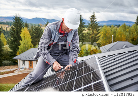 Man builder mounting photovoltaic solar panels on roof of house. Engineer in helmet installing solar module system with help of hex key. Concept of alternative, renewable energy. 112017922