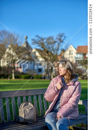 Winter Wonderland Elegance: Beautiful Girl in Pink Jacket Enjoys Festive Atmosphere in Bitigheim-Bissingen Park. girl in a pink winter jacket sits on a bench in a park against the backdrop of the 112018414