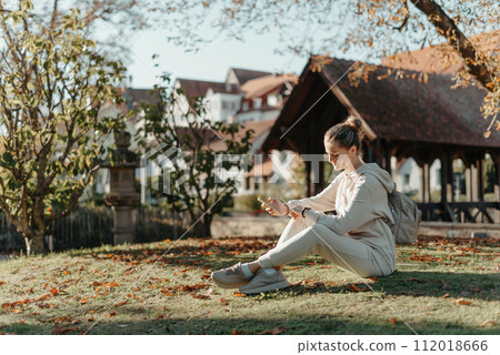 Young Fashionable Teenage Girl With Smartphone In Europian Park In Autumn Sitting At Smiling. Trendy Young Woman In Fall In Park Texting. Retouched, Vibrant Colors. Beautiful Blonde Teenage Girl 112018666