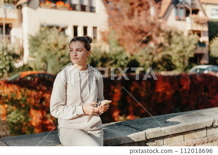 Young Fashionable Teenage Girl With Smartphone In Europian Park In Autumn Sitting At Smiling. Trendy Young Woman In Fall In Park Texting. Retouched, Vibrant Colors. Beautiful Blonde Teenage Girl 112018696