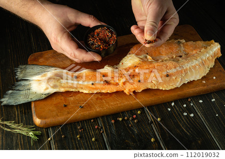 Cooking a fish dish on the kitchen table with the hands of a cook for lunch. Adding spices to fish steak before baking or frying in a pan 112019032