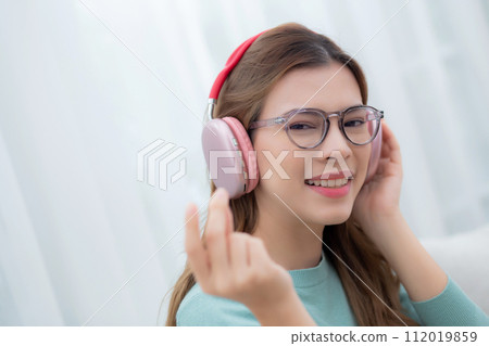 Happy young woman wearing headphones listening music while sitting on sofa. 112019859
