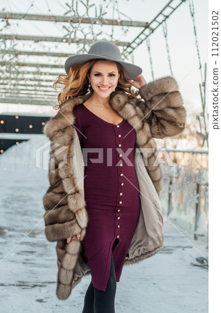 Brunette woman standing on snow city street wearing hat red dress and fur coat 112021502
