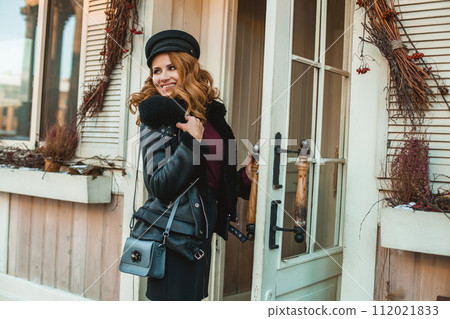 Smiling brunette woman in black leather jacket with blue handbag standing near shop door Smiling brunette woman in black leather jacket with blue handbag standing near shop door 112021833