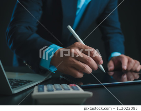 Businessman in black suit working on laptop computer and tablet and calculator, Hand Hands touch on tablet at office with dark background, Online working, Close up, Copy space. Businessman in black suit working on laptop computer and tablet and calculator, Hand Hands touch on tablet at office with dark background, Online working, Close up, Copy space. 112022083