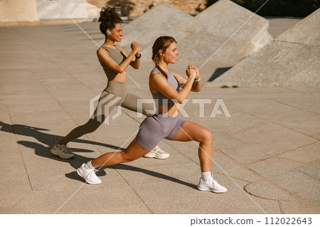 Two young sporty women in tracksuits are stretches legs outside on modern building background 112022643