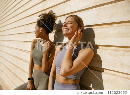 Smiling female sportswomen have a rest after morning jogging outdoors and listen music in headphones Smiling female sportswomen have a rest after morning jogging outdoors and listen music in headphones 112022663