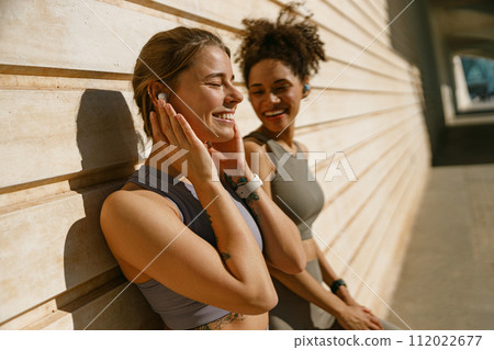 Smiling female sportswomen have a rest after morning jogging outdoors and listen music in headphones 112022677