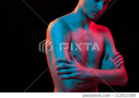 Cropped portrait of bare-chested young handsome man posing crossing hands in neon light against black studio background. 112023580