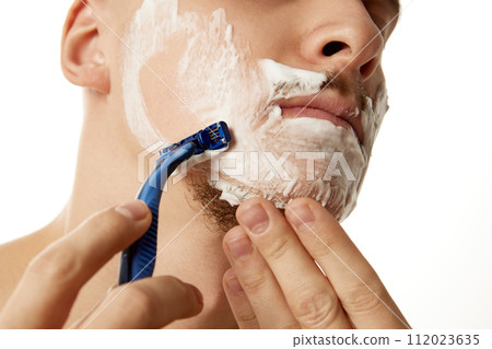 Morning ritual. Close up portrait of young shirtless man carefully shaving face with razor against white studio background. 112023635