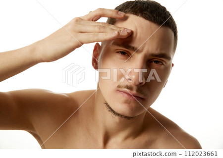 Shirtless young man holding his hand to his forehead and looking to camera with serious expression against white studio background. Shirtless young man holding his hand to his forehead and looking to camera with serious expression against white studio background. 112023661
