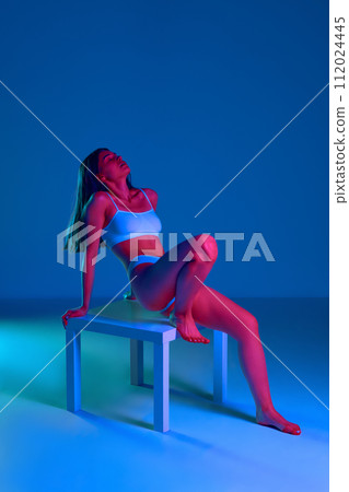 Sensual, tender, young woman with slim, fit body posing on chair in underwear against blue studio background in neon light Sensual, tender, young woman with slim, fit body posing on chair in underwear against blue studio background in neon light 112024445