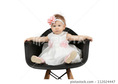 The child is one year old. Beautiful little girl sits on a chair on a white background. Funny child with a bow. Charging baby. The child is one year old. Beautiful little girl sits on a chair on a white background. Funny child with a bow. Charging baby. 112024447