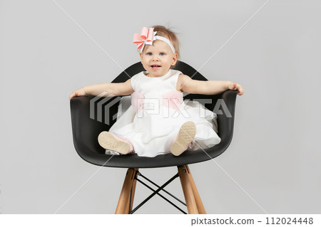The child is one year old. Beautiful little girl is sitting on a chair. Funny child with a bow. Charging baby. The child is one year old. Beautiful little girl is sitting on a chair. Funny child with a bow. Charging baby. 112024448