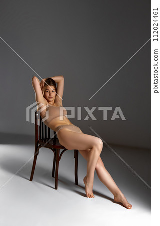 Attractive young slim woman sitting on chair in underwear against gray studio background. Self-love and care 112024461