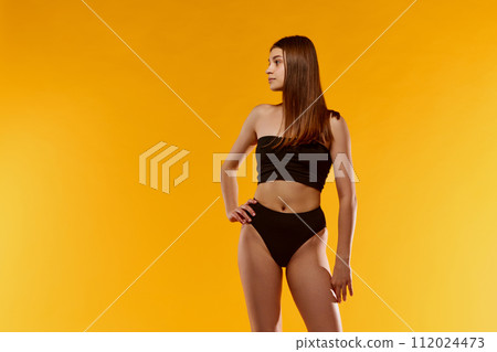 Well-being. Attractive young woman with slim, tanned body standing in black underwear, swimsuit against yellow studio background Well-being. Attractive young woman with slim, tanned body standing in black underwear, swimsuit against yellow studio background 112024473