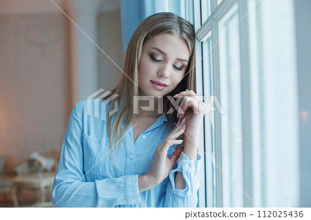 Attractive young woman with long blonde hair wearing blue shirt relaxing at home Attractive young woman with long blonde hair wearing blue shirt relaxing at home 112025436