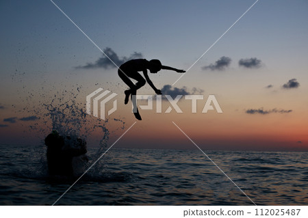Happy holidays concept. Young kid jumping in the sea under sunset sky with sun and cloud 112025487
