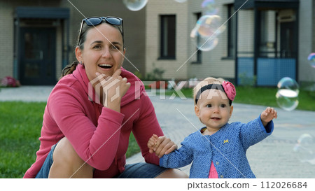 Cute baby girl with her mother watching flying soap bubbles in summer day Cute baby girl with her mother watching flying soap bubbles in summer day 112026684