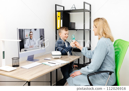 Caucasian little boy making inhalation with nebulizer at home. A young mother helping her son to inhale with a nebulizer at an online meeting with a doctor 112026916