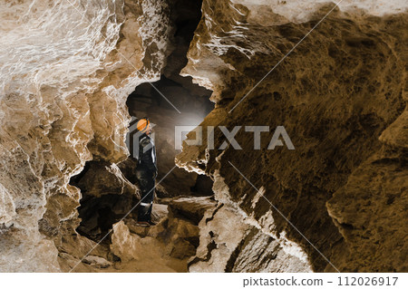 Man walking and exploring dark cave with light headlamp underground. Mysterious deep dark, explorer discovering mystery moody tunnel looking on rock wall inside. 112026917