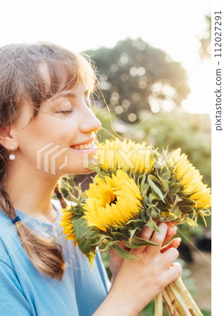Dreaming young female farmer woman with closed eyes holding and sniffing a sunflowers bouquet on the green garden background in sunset light. Enjoy the moment. Rural, Cottage core lifestyle. Vertical Dreaming young female farmer woman with closed eyes holding and sniffing a sunflowers bouquet on the green garden background in sunset light. Enjoy the moment. Rural, Cottage core lifestyle. Vertical 112027291