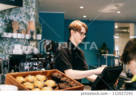Young waiter serving customer at cash point in cafe. Man working with POS terminal. Cashier, barista checking for payment receipt. Hospitality, server and preparing a slip at the till in coffee shop. 112027293