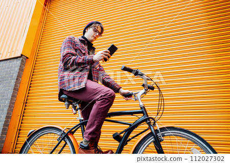 Young hipster man using mobile phone while sitting on cruiser bicycle near orange wall. Student using 5g network,rests after riding. Eco friendly transport for carbon footprint. Sustainable lifestyle 112027302