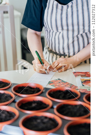 No face woman making notes on stickers for plant pots with planted seeds at home kitchen. Preparing for new kitchen garden season. Sowing seeds. Vertical card. Soft selective focus, copy space. 112027312