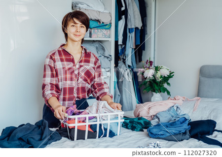 Smiling Woman with basket of selected clothes from her wardrobe for donating to a Charity. Decluttering, Sorting clothes And Cleaning Up. Reuse, second-hand concept. Conscious consumer, sustainability 112027320