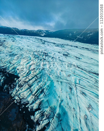 Aerial view of vatnajokull glacier mass in iceland, beautiful ice rocks with massive crevasse around snowy hills and scenery. Majestic frozen icebergs floating on freezing cold lake. Aerial view of vatnajokull glacier mass in iceland, beautiful ice rocks with massive crevasse around snowy hills and scenery. Majestic frozen icebergs floating on freezing cold lake. 112031088